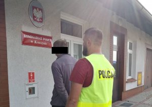 Zatrzymany mężczyzna i policjant w kamizelce z napisem POLICJA stojący przed wejściem do Komisariatu Policji w Kruszynie.