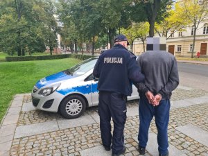 Umundurowany policjant prowadzący zatrzymanego mężczyznę, w tle radiowóz.