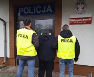 Dwóch policjantów w kamizelkach odblaskowych prowadzących zatrzymanego mężczyznę, w tle budynek Komisariatu Policji w Kruszynie.
