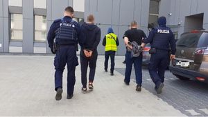 Na zdjęciu widać tyłem 5 idących mężczyzn, po zewnętrznej dwóch umundurowanych policjantów w środku policjant w kamizelce z napisem POLICJA i dwóch zatrzymanych z kajdanami założonymi na ręce trzymane z tyłu. W tle budynek, po prawej stronie dwa pojazdy.