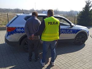 Policjant kamizelce z napisem POLICJA stoi tyłem razem z zatrzymanym, który ma kajdanki założone na ręce trzymane  z tyłu. Przed nimi radiowóz.