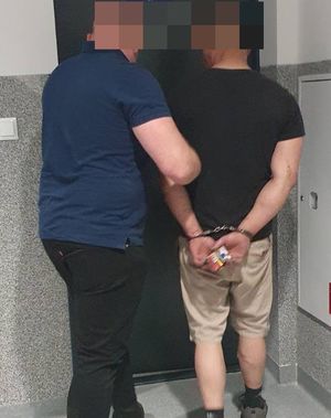 Policjant kryminalny stoi przed drzwiami z zatrzymanym mężczyzną, który ma założone kajdanki na rękach trzymanych z tyłu.