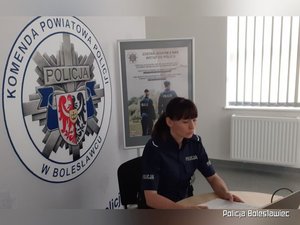 Policjantka siedząca przed komputerem, za jej plecami widoczny baner z logo Komenda Powiatowa Policji w Bolesławcu.