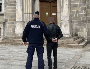 Umundurowany policjant i zatrzymany mężczyzna z kajdankami założonymi na ręce trzymane z tyłu, stoją na tle budynku.