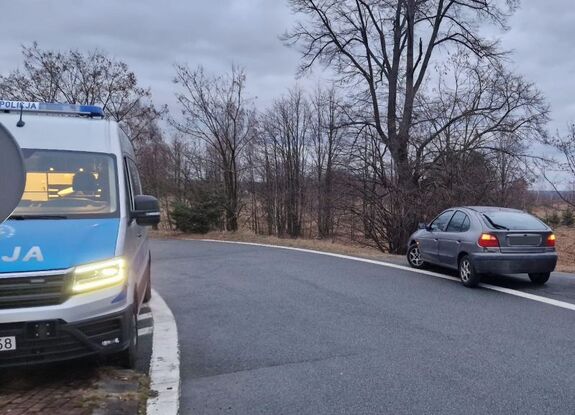 Dwa pojazdy stojące po dwóch stronach jezdni. Po prawej szary samochód osobowy, po lewej oznakowany policyjny bus.