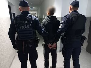 Umundurowani policjanci prowadzą mężczyznę z kajdankami założonymi na ręce trzymane z tyłu.