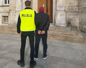 Policjant w kamizelce odblaskowej z napisem POLICJA prowadzący zatrzymanego mężczyznę z kajdankami założonymi na ręce trzymane z tyłu. W tle budynek sądu.