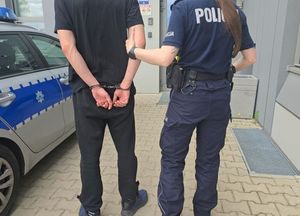 Mężczyzna z kajdankami założonymi na ręce trzymane z tyłu prowadzony przez umundurowaną policjantkę.