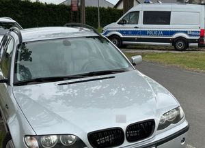 Na pierwszym planie po lewej stronie widoczny przód srebrnego pojazdu osobowego, w tle po prawej stronie radiowóz typu bus.