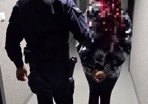 Umundurowany policjant prowadzący kobietę z kajdankami założonymi na ręce trzymane z tyłu, górna cześć wizerunku podejrzanej jest wypikselowana. Kobieta po prawej stronie, a policjant po lewej.