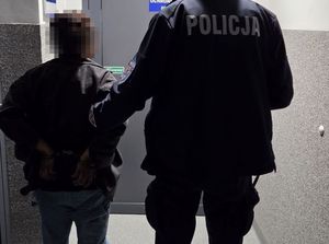 Umundurowany policjant prowadzący kobietę z kajdankami założonymi na ręce trzymane z tyłu, górna cześć wizerunku podejrzanej jest wypikselowana. Kobieta po lewej stronie a policjant po prawej.