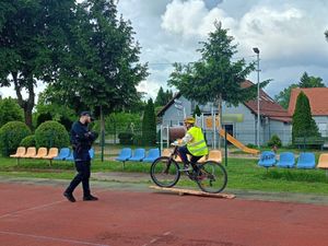 Uczeń jadący na rowerze w kasku ochronnym i kamizelce odblaskowej oraz stojący obok umundurowany policjant.