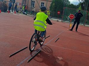 Na pierwszym planie uczeń jadący na rowerze w kasku ochronnym i kamizelce odblaskowej oraz stojący przed nim umundurowany policjant.