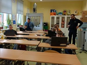 Uczniowie w klasie siedzący w ławkach przed monitorami oraz stojący po prawej stronie umundurowany policjant oraz  w tylnej części klasy nauczyciel.