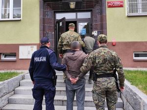 Na pierwszym planie widoczny policjant i funkcjonariusz Straży Granicznej prowadzący zatrzymanego mężczyznę , który ma założone kajdanki na ręce trzymane z tyłu. Na drugi planie widoczny inny zatrzymany prowadzony przez funkcjonariusza Straży Granicznej. W tle budynek, siedziba Straży Granicznej w Zgorzelcu.