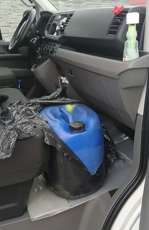 Niebieski kanister częściowo okryty czarna folią znajdujący się na podłodze busa, pomiędzy fotelem pasażera i pulpitem.