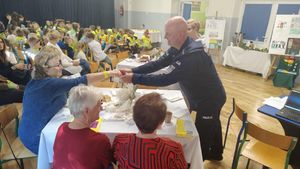 Policjant podający element odblaskowy seniorkom siedzącym przy stole, w tle widoczne dzieci w zółtych odblaskowych kamizelkach.