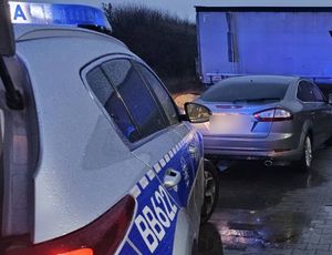 Na pierwszym planie widoczny prawy bok radiowozu, dalej widać pojazd osobowy z zamazanymi tablicami rejestracyjnymi.
