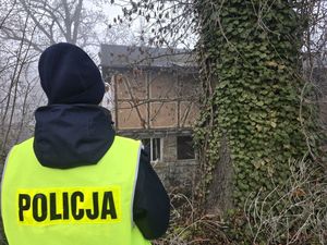 Na zdjęciu widoczna stojąca tyłem policjantka w żółtej kamizelce z napisem policja, w tle budynek bez okien.