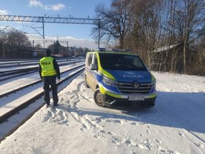 Umundurowany policjant stojący obok radiowozu wzdłuż torowiska, w tle widoczne budynki.
