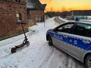 Na zdjęciu widoczny jest radiowóz po prawej stronie, hulajnoga po lewej stronie na tle budynków. Na drugim planie widoczna jezdnia.