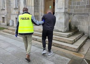 Policjant w żółtej kamizelce z napisem POLICJA prowadzi zatrzymanego mężczyznę. W tle budynek sądu.