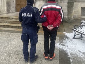 Umundurowany policjant prowadzący mężczyznę z kajdankami założonymi na ręce trzymane  tyłu, w tle budynek.