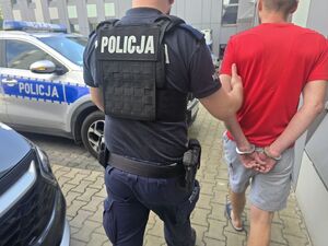 Umundurowany policjant prowadzi mężczyznę w czerwonej koszulce, który ma założone kajdanki na ręce trzymane z tyłu.