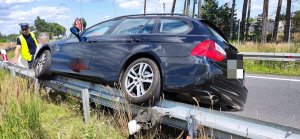 Na zdjęciu widać czarne auto, które swoją lewą częścią najechało na barię energochłonną, z przodu pojazdu stoi umundurowany policjant.